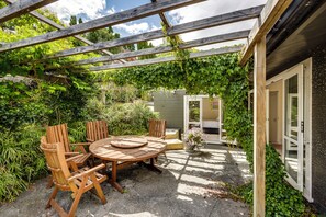 Een houten tafel met stoelen staat buiten onder een houten pergola, omringd door weelderige begroeiing en een klein gebouw met een deur.