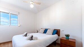 Un dormitorio con cama, mesita de noche, ventilador de techo, ventana y una planta pequeña.