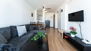 Una sala de estar moderna con un sofá gris, una mesa de centro con una planta, una zona de comedor con mesa y sillas, un televisor montado en la pared y un ventilador de techo.