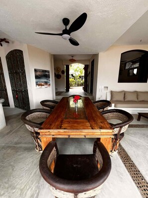 A dining area with a long wooden table, wicker chairs, a ceiling fan, and a view of the outdoors. - Quaint 3-bedroom house with WiFi in Troncones (Troncones)