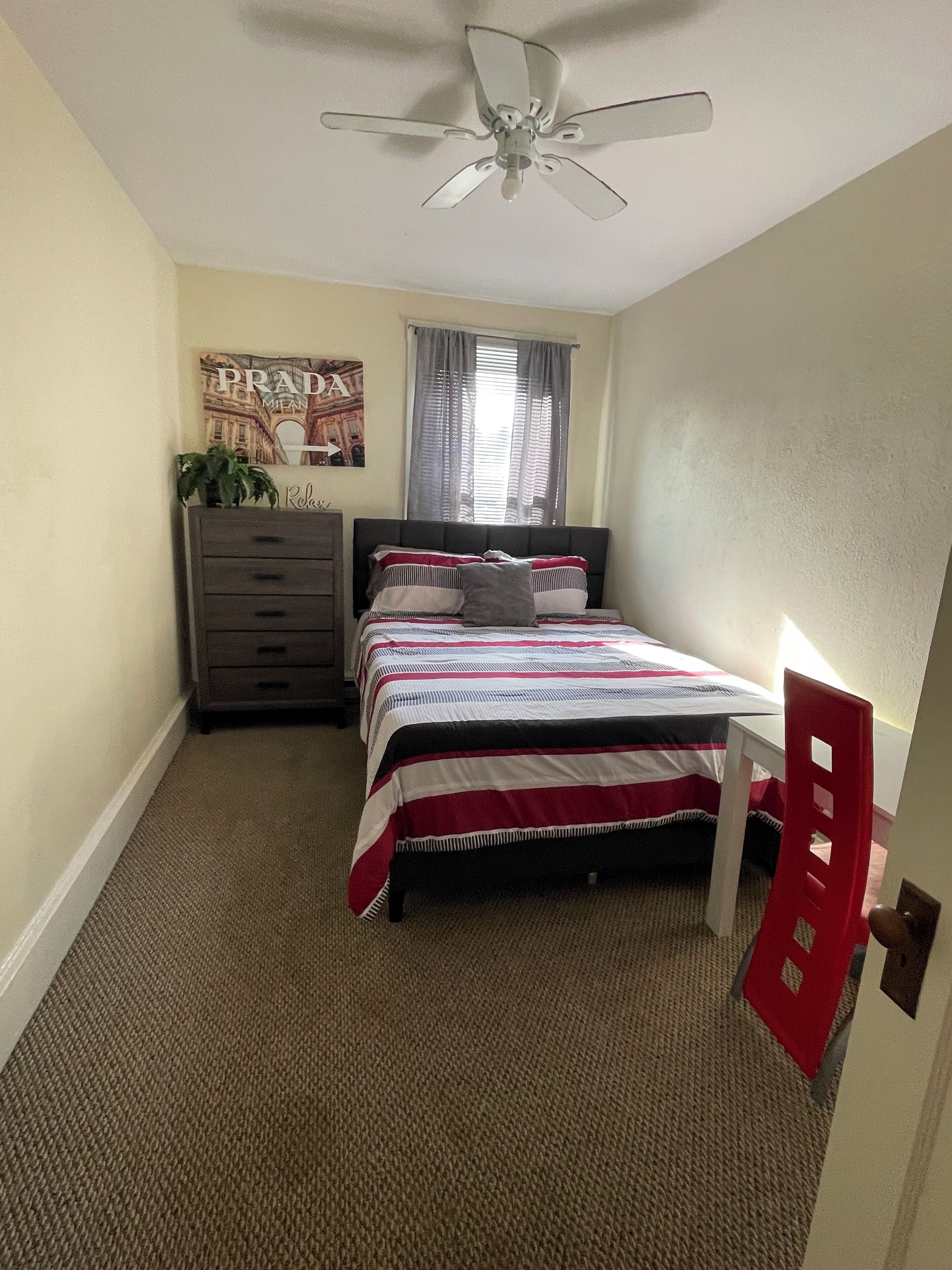 A bedroom with a bed, a dresser, a ceiling fan, a window with curtains, and a door.
