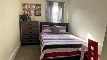 A bedroom with a bed, a dresser, a ceiling fan, a window with curtains, and a door.