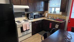A compact kitchen with dark wooden cabinets, a black refrigerator, an oven, a microwave, and a sink. There is a dining table with chairs and a window with blinds.