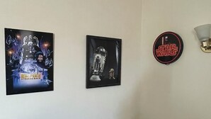 A room with a white wall, a framed picture of a Star Wars character, a framed picture of a robot, and a Star Wars-themed light fixture.