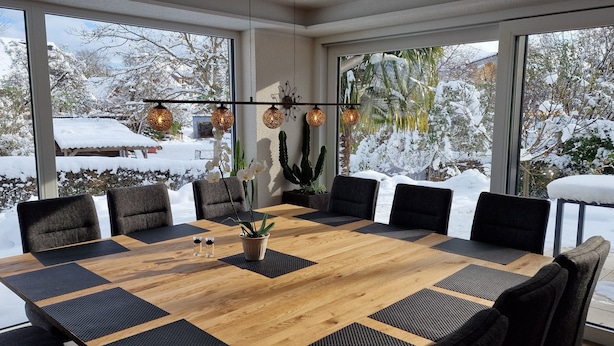 Un comedor moderno con una amplia mesa de madera rodeada de sillas, grandes ventanales con vistas a un paisaje nevado y una original lámpara de techo.