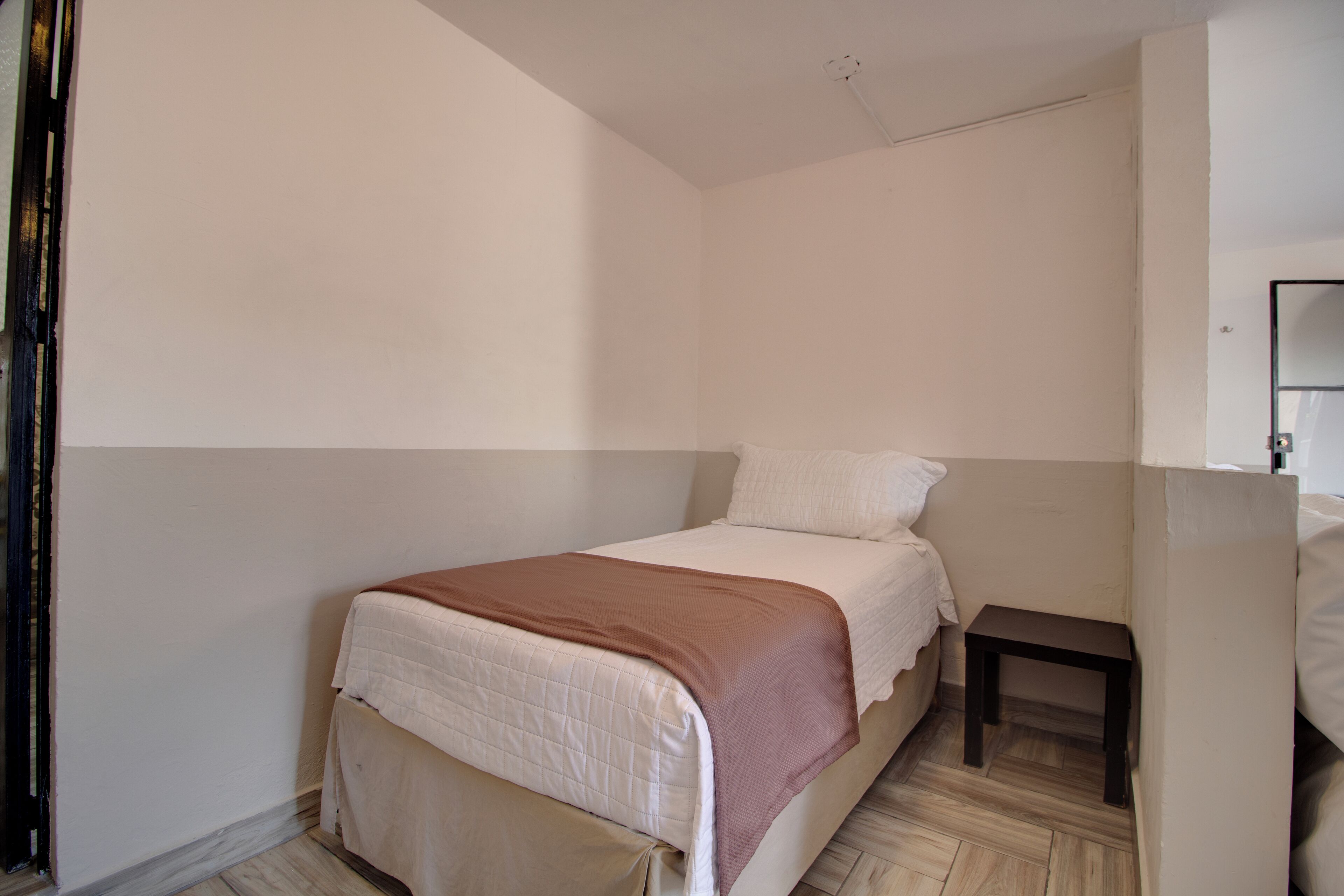 Une petite chambre avec un lit simple, un plancher en bois, une table de chevet et un mur blanc.