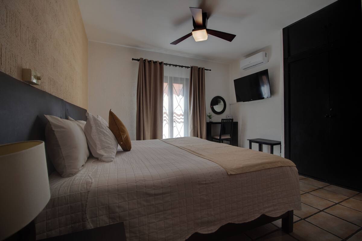 Une chambre à coucher avec un lit, un ventilateur de plafond, une télévision et une fenêtre avec des rideaux.