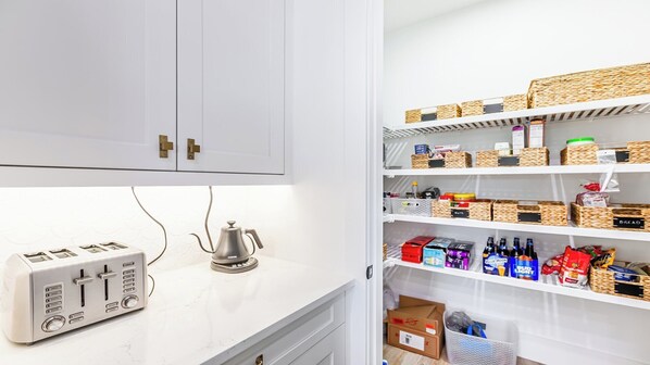 A kitchen with a built-in toaster, a kettle, and shelves stocked with various food items and kitchen supplies.