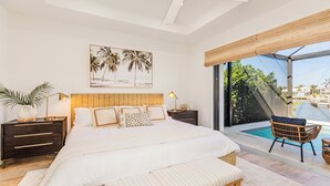 Ein Schlafzimmer mit einem großen Bett, einem Nachttisch, einem Beistelltisch, einem Fenster mit Blick auf einen Pool und Palmen sowie einem Wandbild.