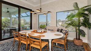 Un espace repas avec une table ronde, des chaises en bois, un lustre et de grandes fenêtres donnant sur l’extérieur.