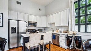 A modern kitchen with white cabinets, stainless steel appliances, a dining area with two chairs, and a view of trees through large windows. - The Retreat off Magazine (New Orleans)