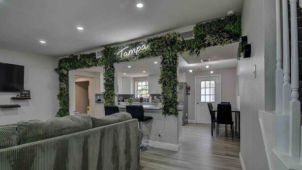 Ein modernes Wohnzimmer mit einem grünen Pflanzenbogen mit dem Schriftzug "Tampa", einem grauen Sofa und einer Küchenzeile mit Esstisch und Stühlen.