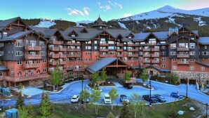 Un lodge de varios pisos con balcones, una pista de esquí al fondo y un área de estacionamiento.