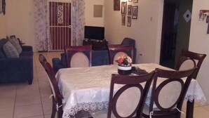 Un comedor con una mesa preparada para cuatro personas, rodeada de sillas de madera. Hay un ventilador de techo sobre la mesa, y un televisor montado en la pared detrás del comedor.