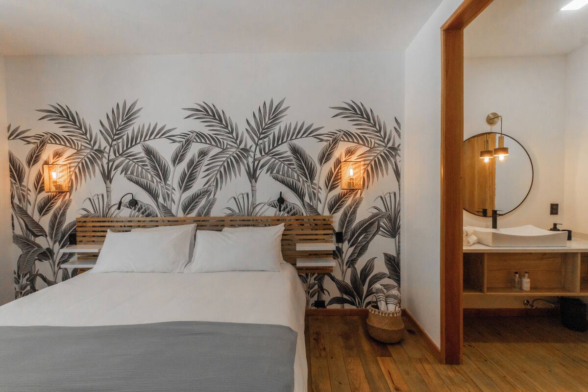 A bedroom with a bed, a wooden headboard, a wall with a tropical leaf pattern, and a bathroom with a wooden vanity and mirror.