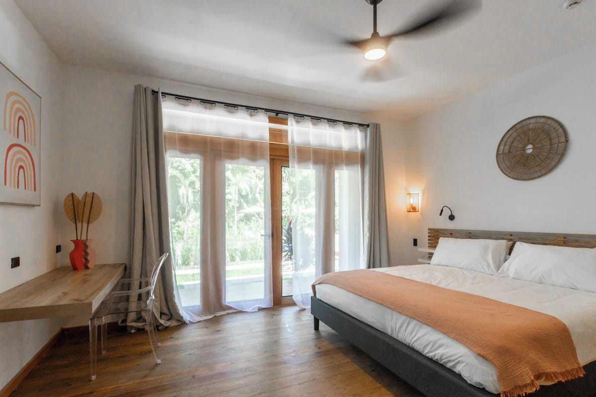 A bedroom with a bed, a desk, and a chair. There is a window with curtains, a ceiling fan, and a wall clock.