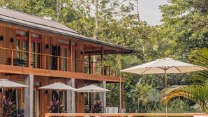 Ein Holzhaus mit Balkon, Außenbereich mit Sonnenschirmen und einem Pool, der von tropischer Vegetation umgeben ist.