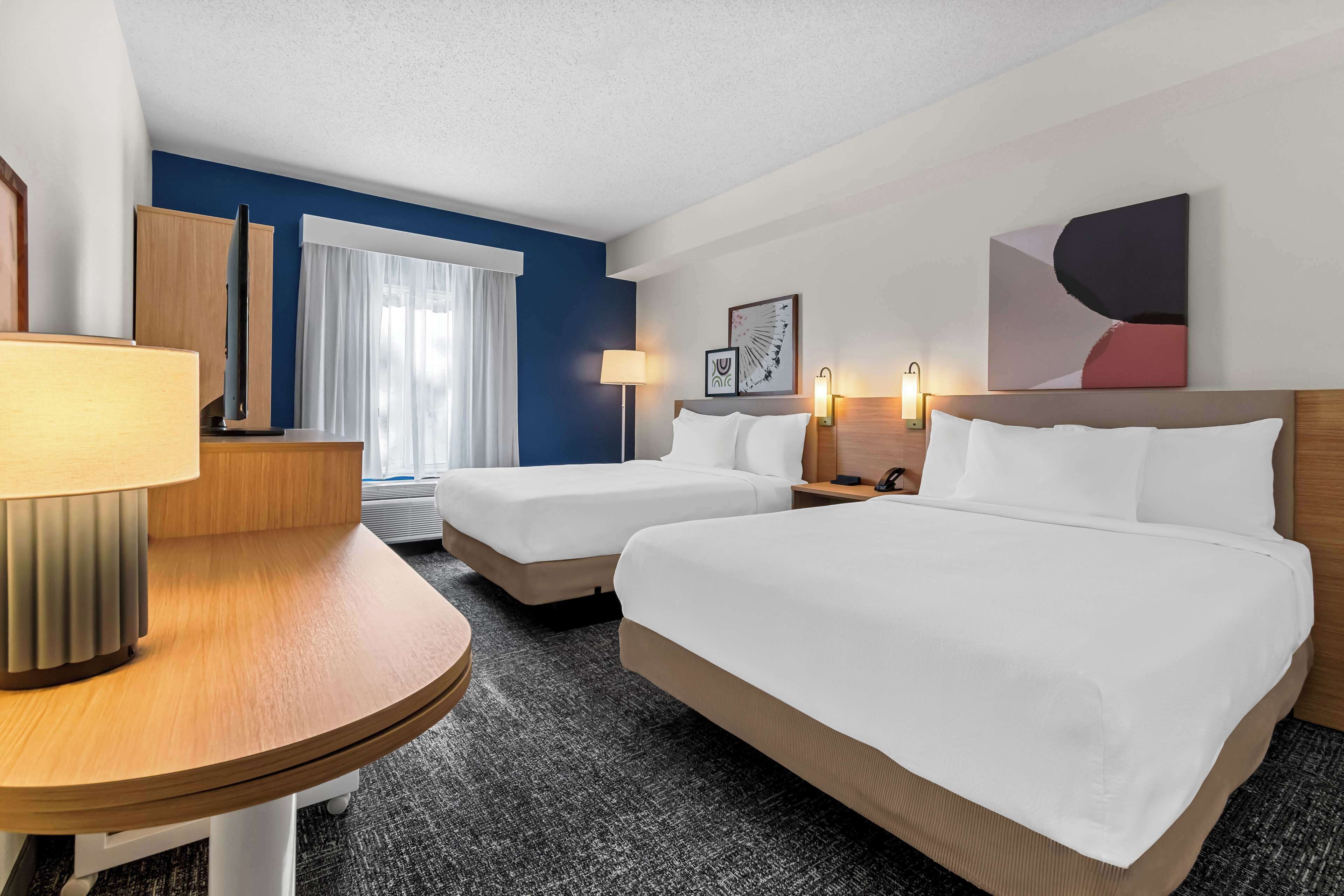 A hotel room with two beds, a desk, a lamp, a flat-screen TV, and a window with curtains.