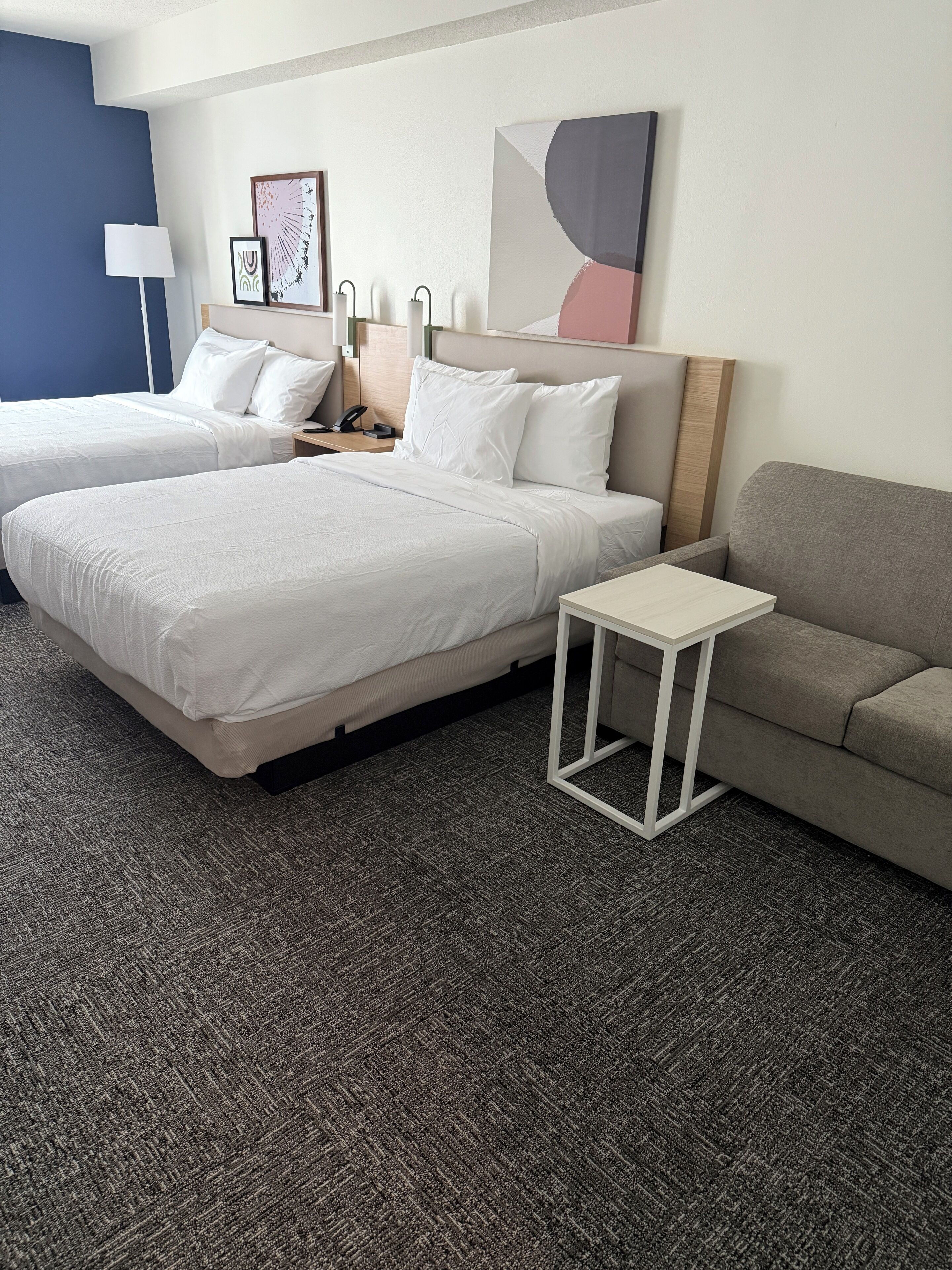 A hotel room with two beds, a grey sofa, a small white table, and a lamp.