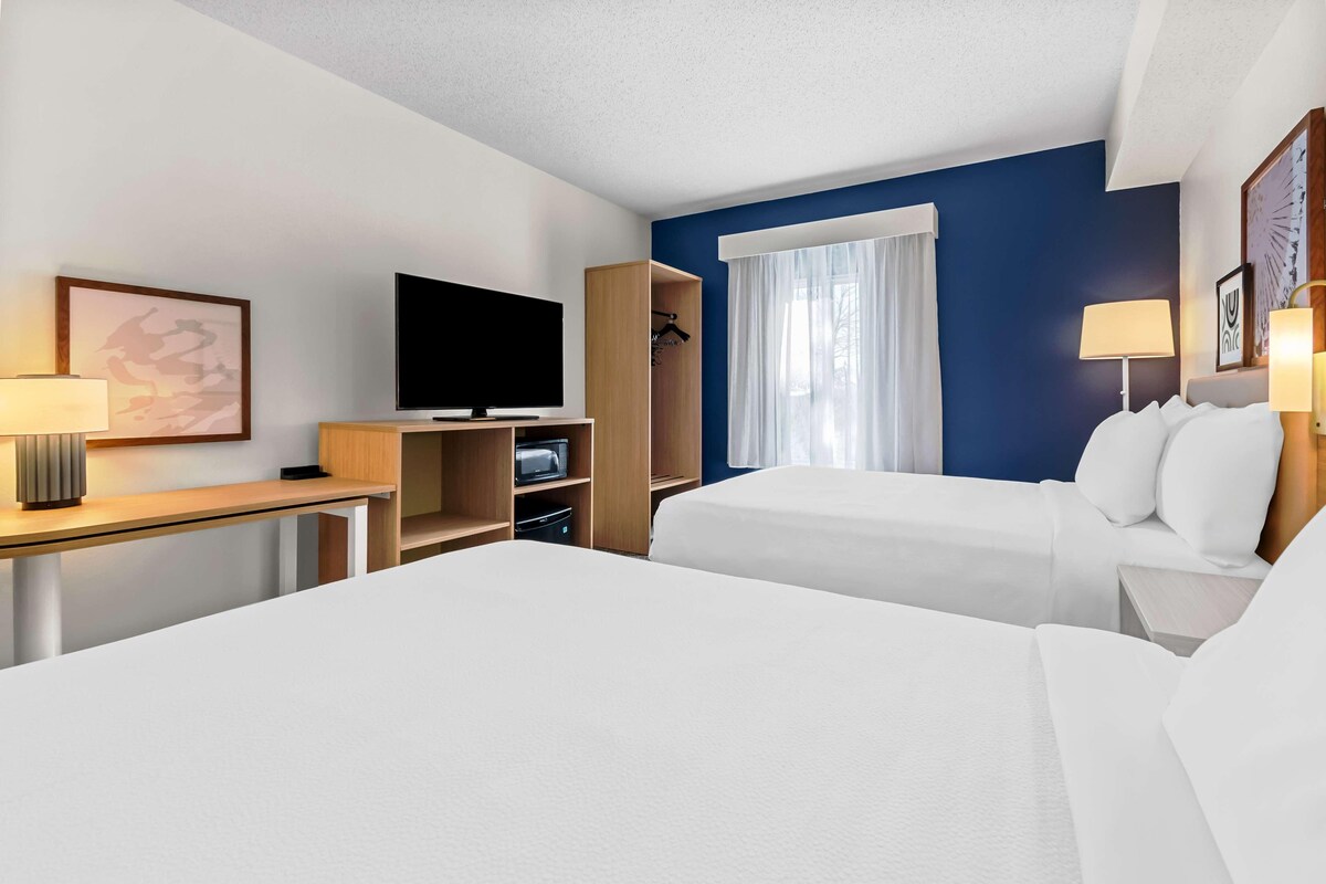 Habitación de hotel con dos camas, televisión, escritorio con lámpara, armario y ventana con cortinas.