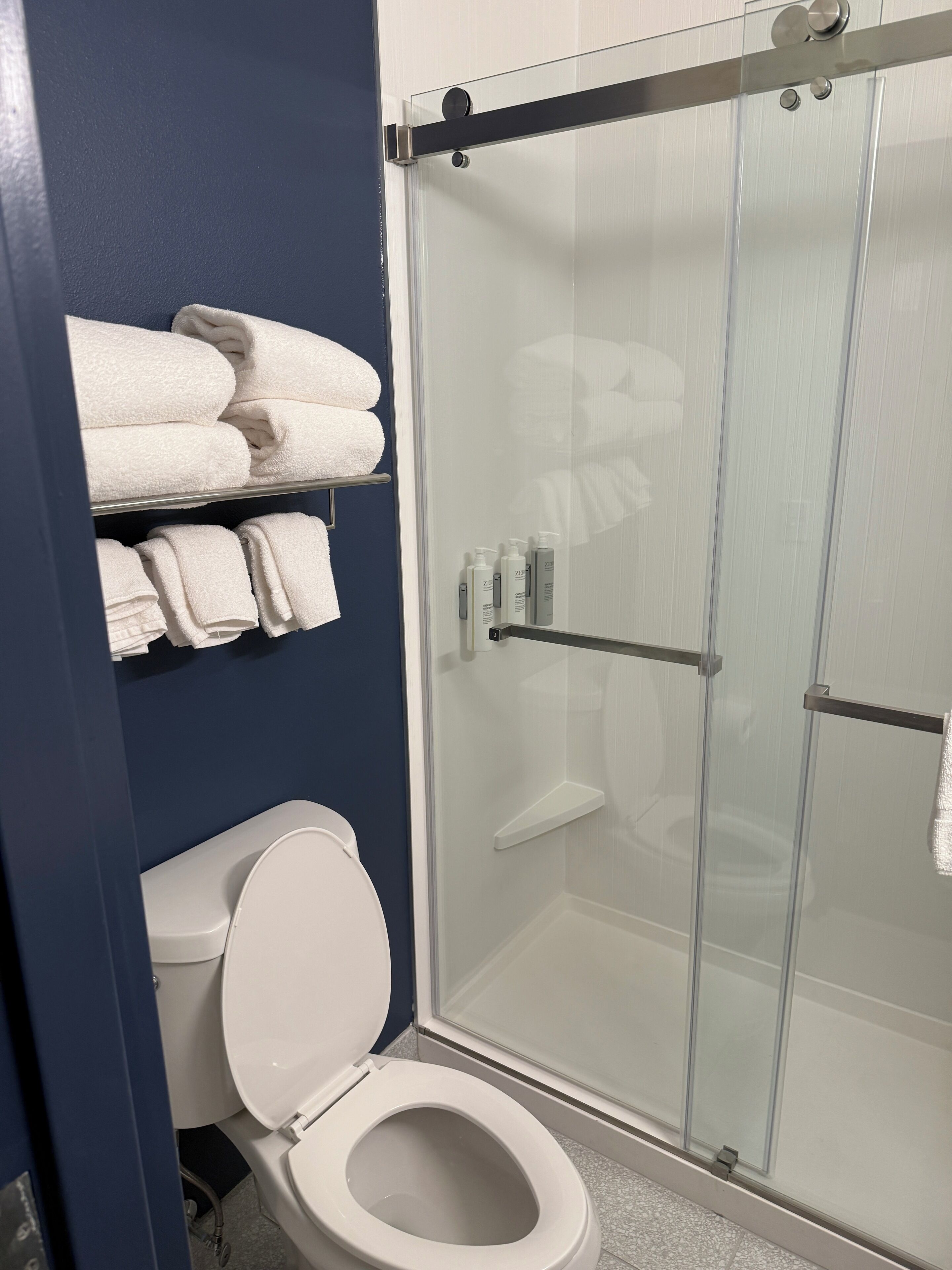 Un baño con inodoro blanco, cabina de ducha de cristal y un perchero con toallas dobladas.
