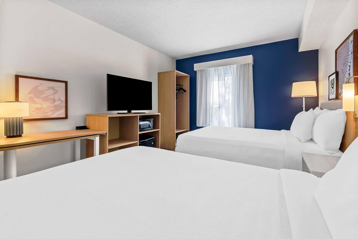 A hotel room with two beds, a TV, a desk with a lamp, a wardrobe, and a window with curtains.