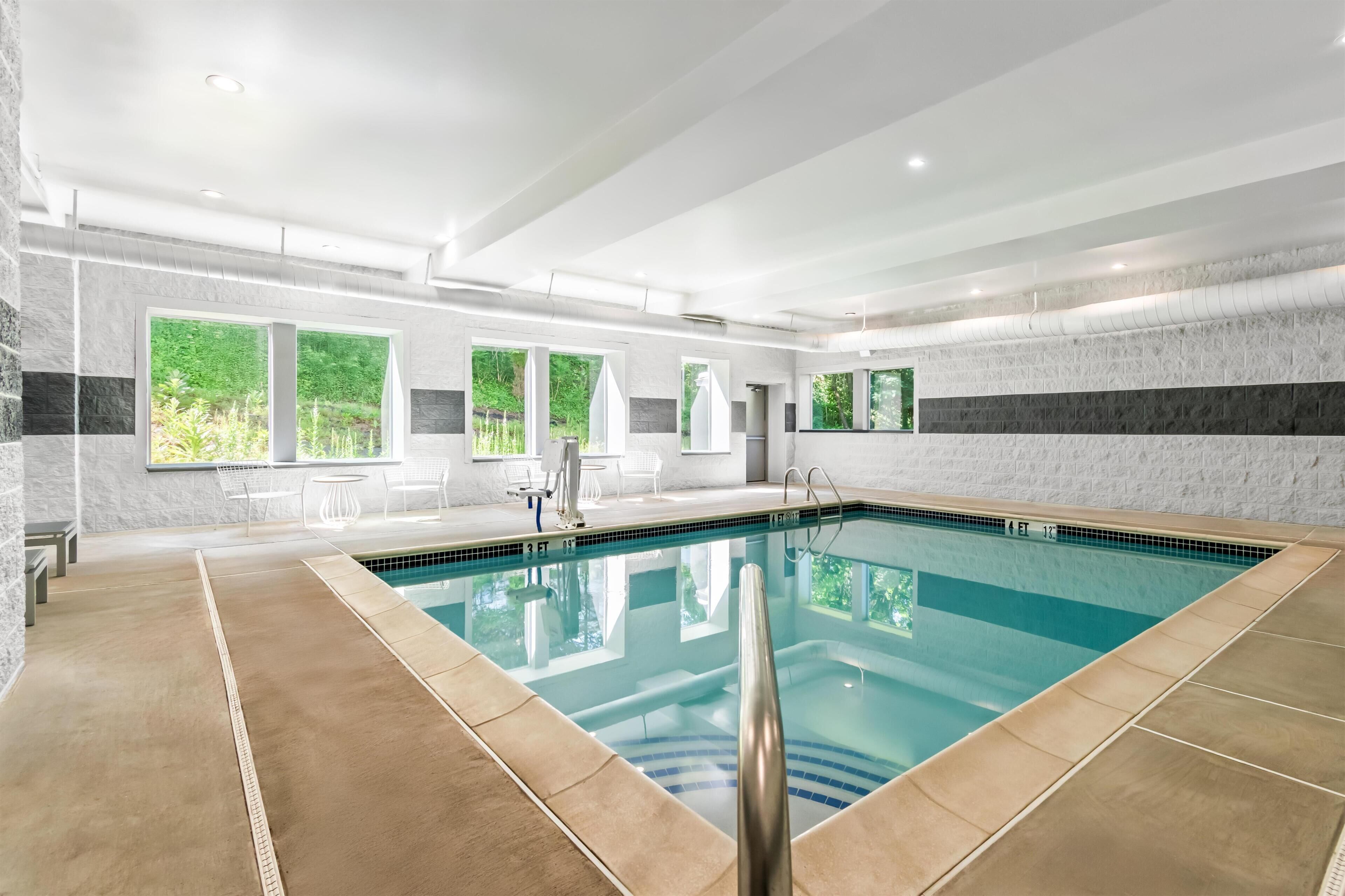 A modern indoor swimming pool with clear water, surrounded by a tiled floor and walls, with windows offering views of greenery outside.