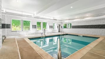 A modern indoor swimming pool with clear water, surrounded by a tiled floor and walls, with windows offering views of greenery outside.