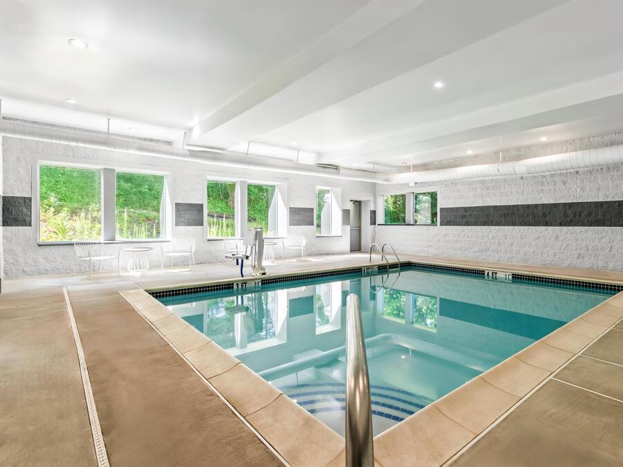 A modern indoor swimming pool with clear water, surrounded by a tiled floor and walls, with windows offering views of greenery outside.