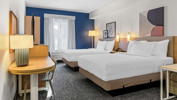 Una habitación de hotel con dos camas, un escritorio, una silla, una lámpara y un ventanal con cortinas.