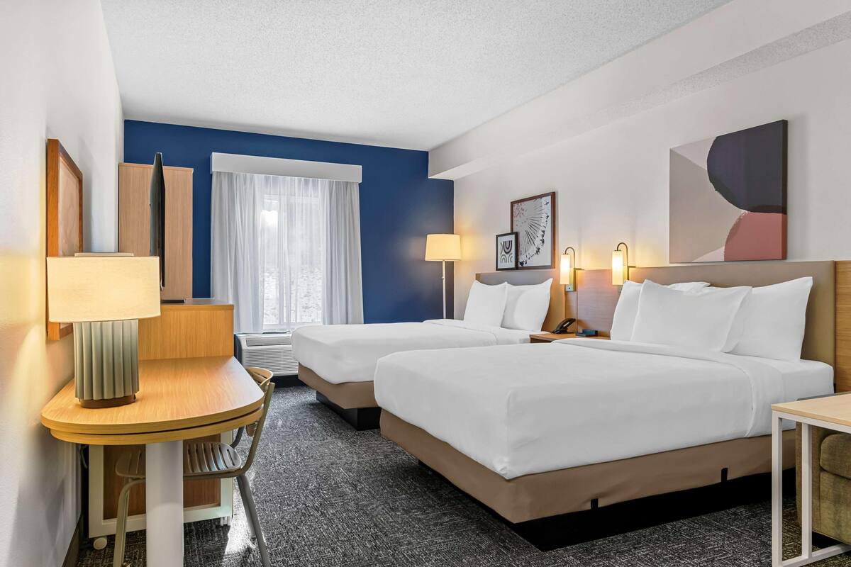 A hotel room with two beds, a desk, a chair, a lamp, a window with curtains, and a wall-mounted artwork.