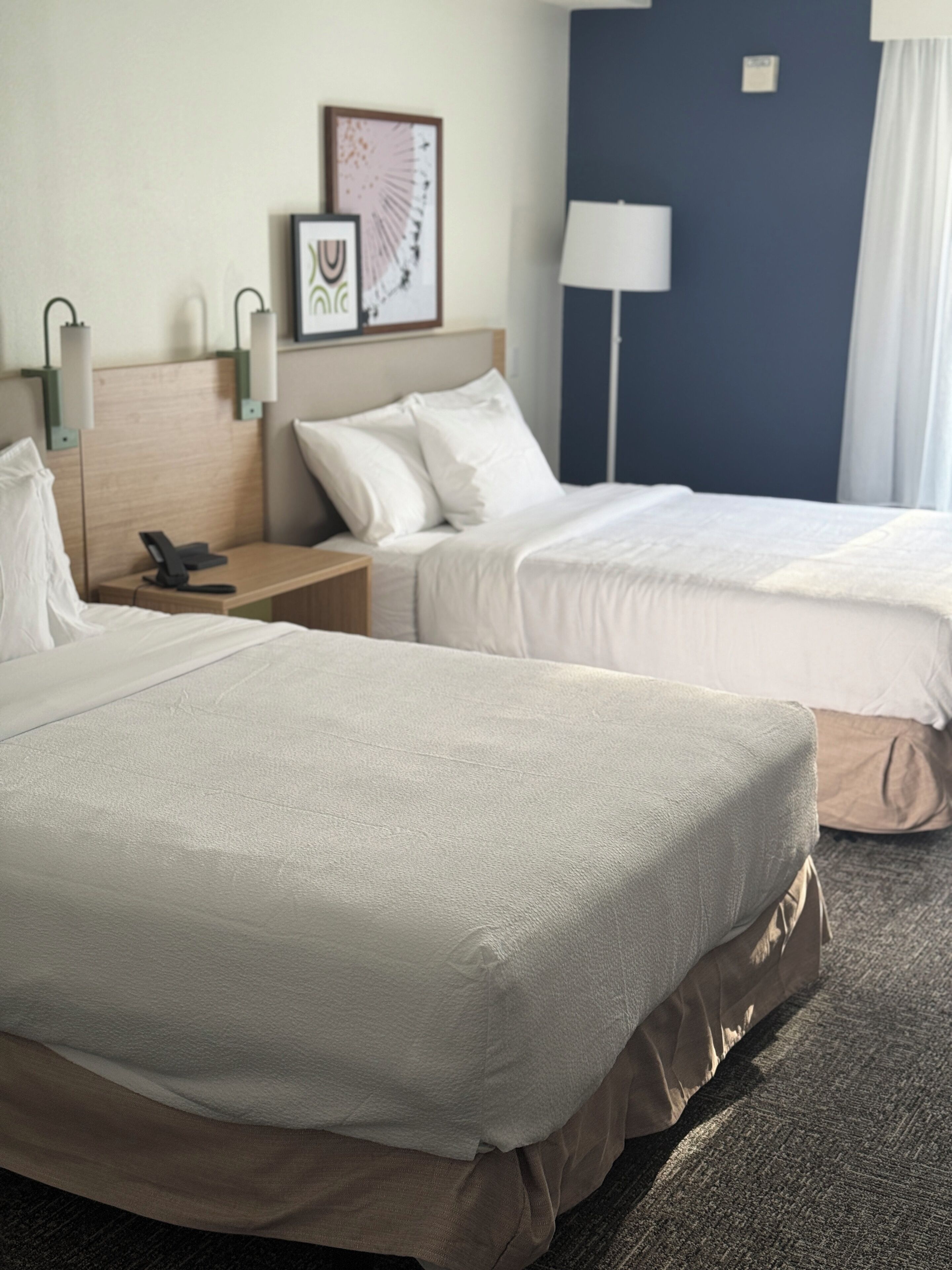 A hotel room with two beds, a bedside table with a phone, a lamp, and a framed picture on the wall.