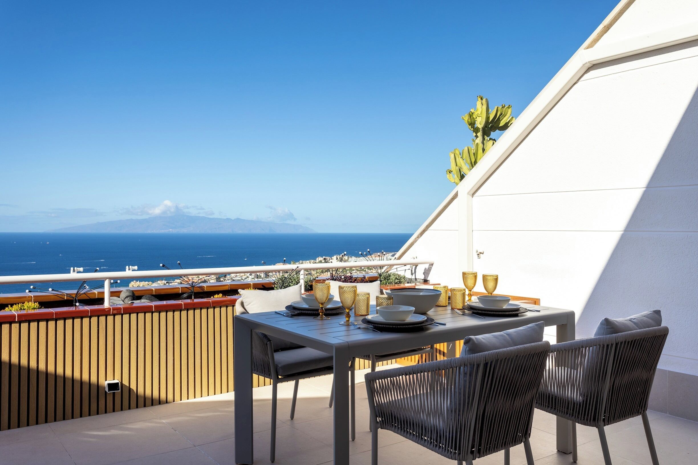 A balcony with a table set for two, overlooking a scenic view of the sea and mountains.