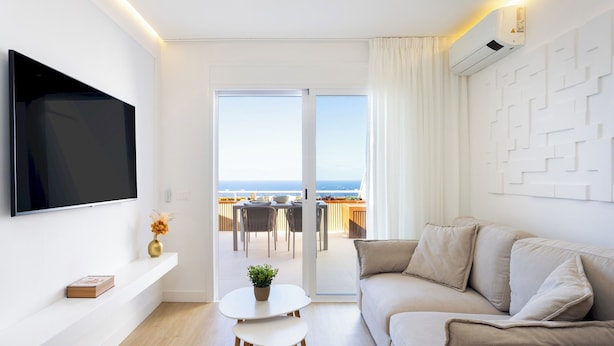A modern living room with a flat-screen TV, a sofa, a small table with a plant, and a balcony with a view of the sea. - Home2Book Costa Adeje Escape & Pool, Only Adults (Adeje)