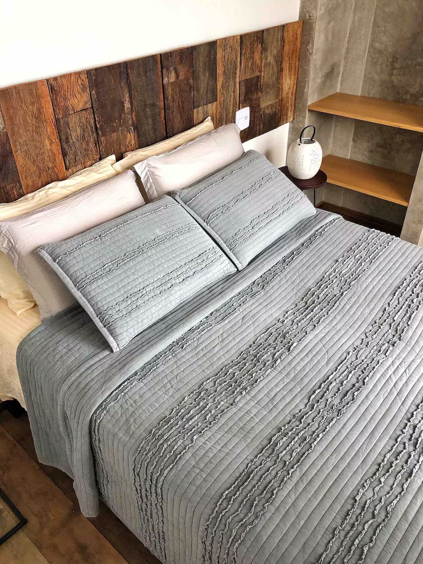 A bed with a grey quilted bedspread and pillows, a wooden headboard, a bedside table with a white lamp, and a shelf.