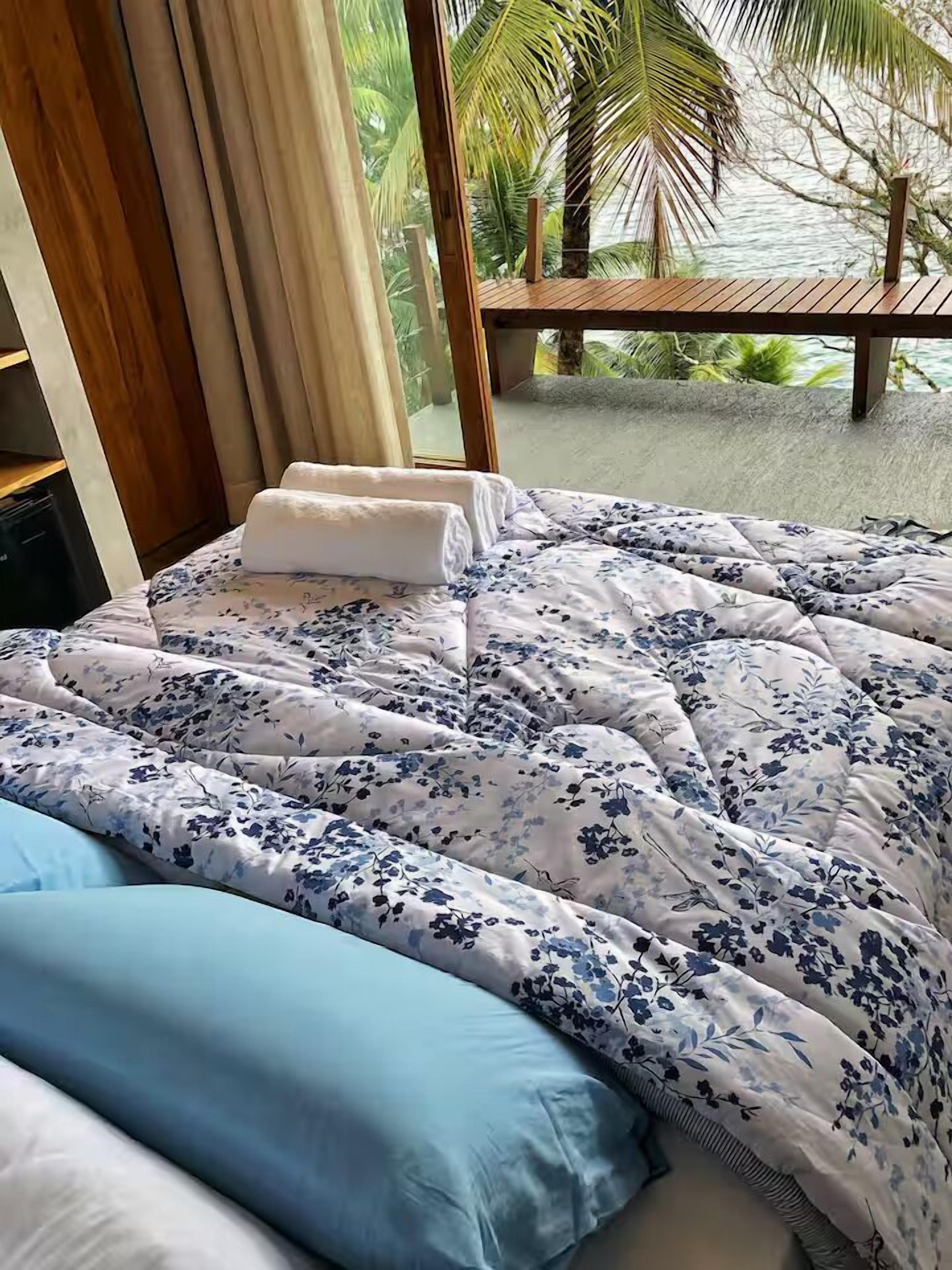 A bed with a floral comforter and a blue pillow, a wooden headboard, and a view of palm trees and a beach through a window.