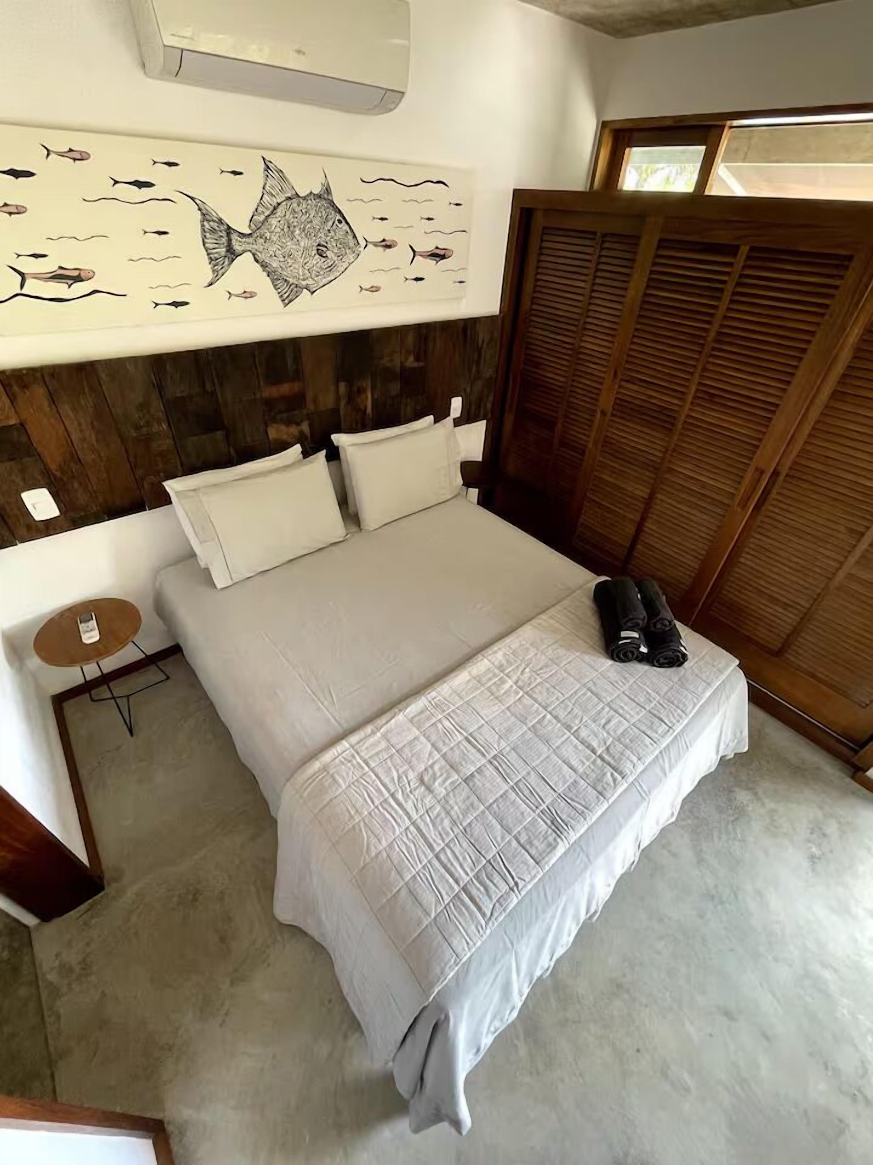 Een slaapkamer met een bed, kussens, een nachtkastje, een schilderij van een vis en een houten garderobe.