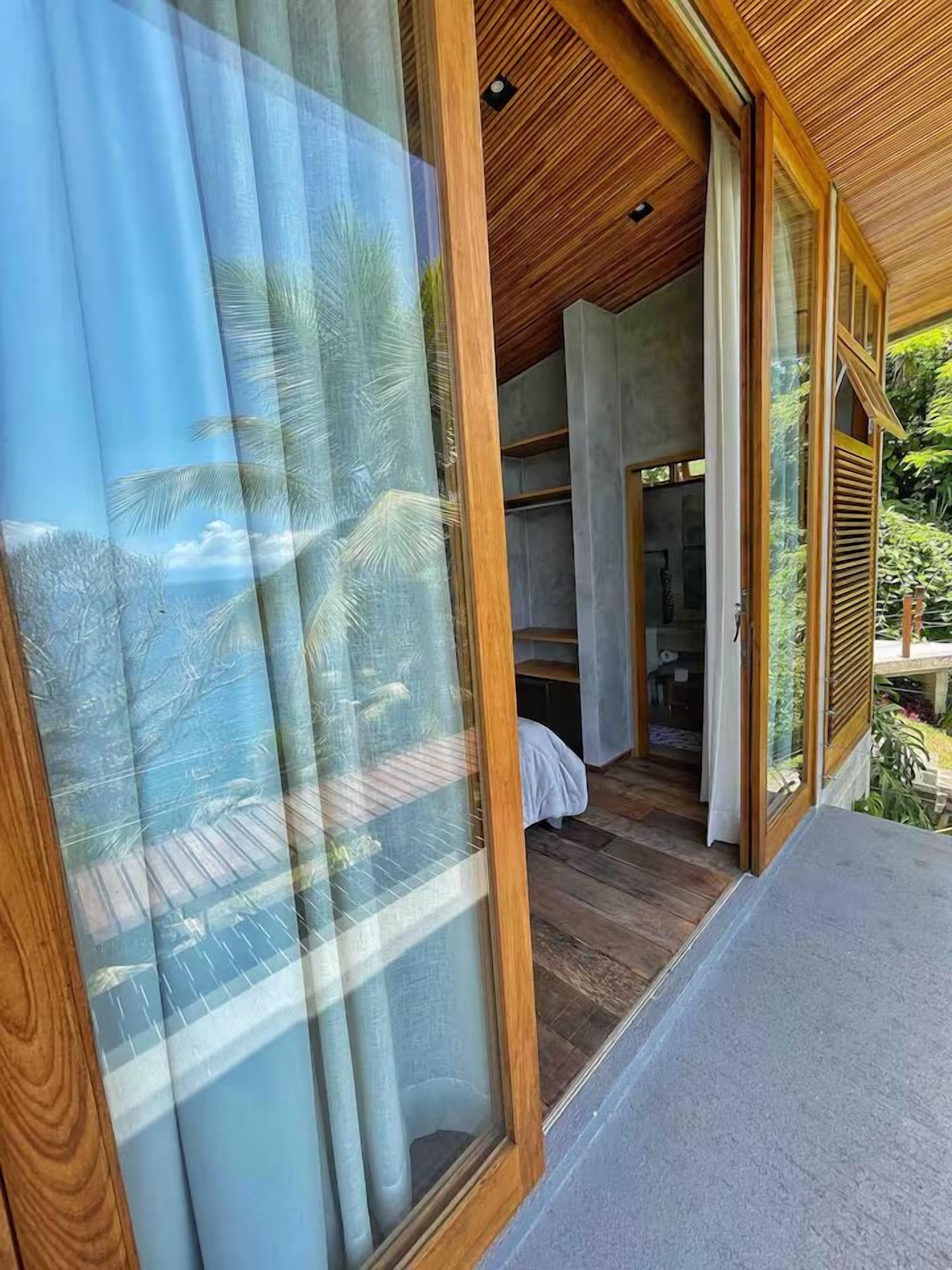 Une chambre moderne dotée de grandes portes-fenêtres coulissantes, d’un plafond en bois et offrant une vue sur un paysage tropical.