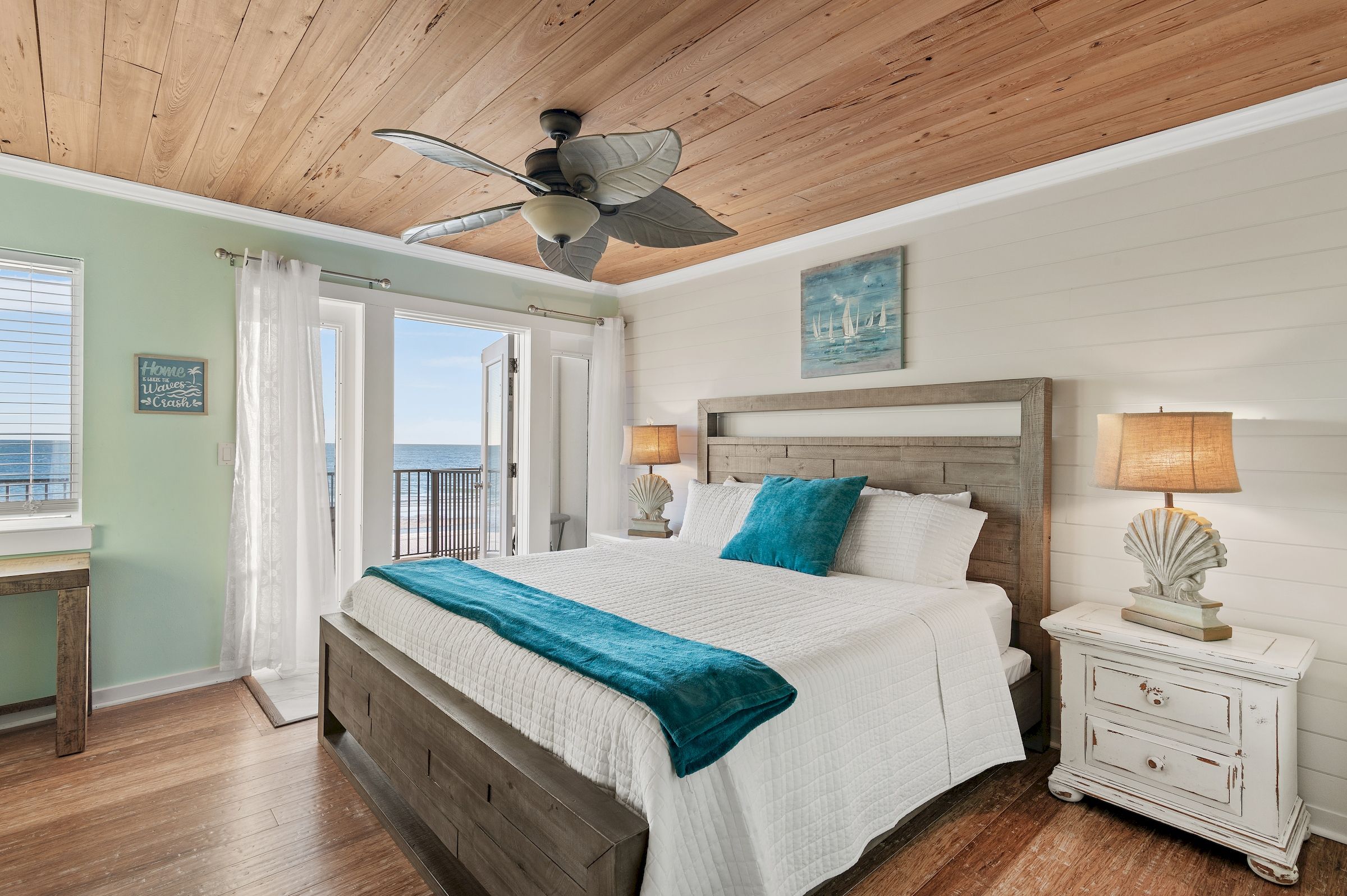 Un dormitorio con techo de madera, cama con colcha blanca y azul turquesa, mesita de noche con lámpara y vistas al exterior a través de una puerta.
