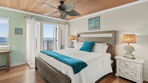 A bedroom with a wooden ceiling, a bed with a white and teal bedspread, a nightstand with a lamp, and a view of the outdoors through a door.