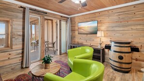 A wooden interior with a ceiling fan, a large window, a green chair, a small table with a plant, and a wooden barrel.