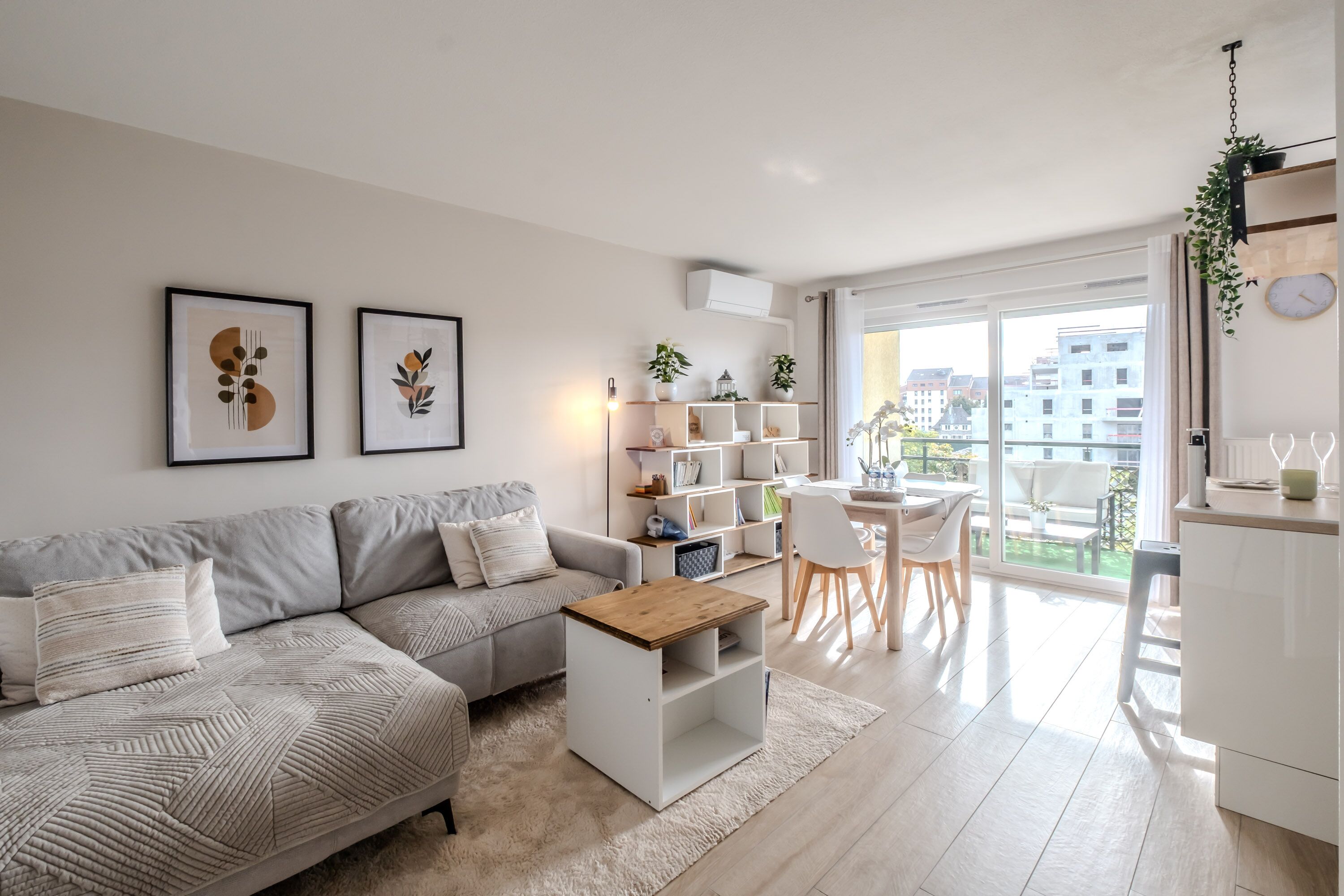 Ein modernes Wohnzimmer mit einem grauen Sofa, einem Couchtisch und einem Essbereich mit Tisch und Stühlen. An der Wand hängen zwei gerahmte Kunstwerke, eine weiße Regaleinheit mit Pflanzen und ein Blick auf Gebäude durch das große Fenster.