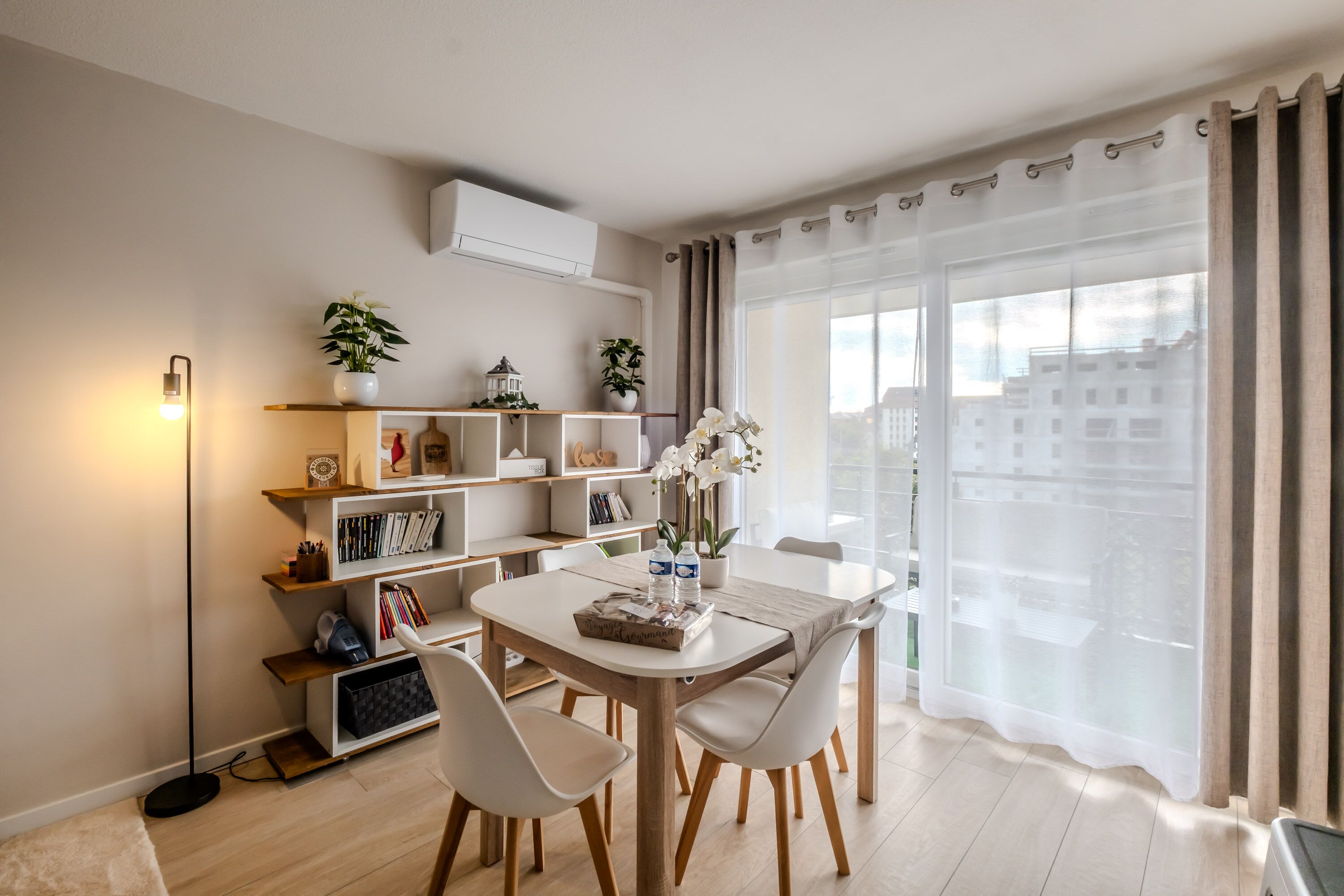 Una sala de estar moderna con una mesa de comedor, sillas blancas, una estantería y un amplio ventanal con cortinas.