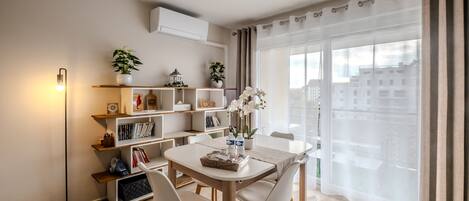 Una sala de estar moderna con una mesa de comedor, sillas blancas, una estantería y un amplio ventanal con cortinas.