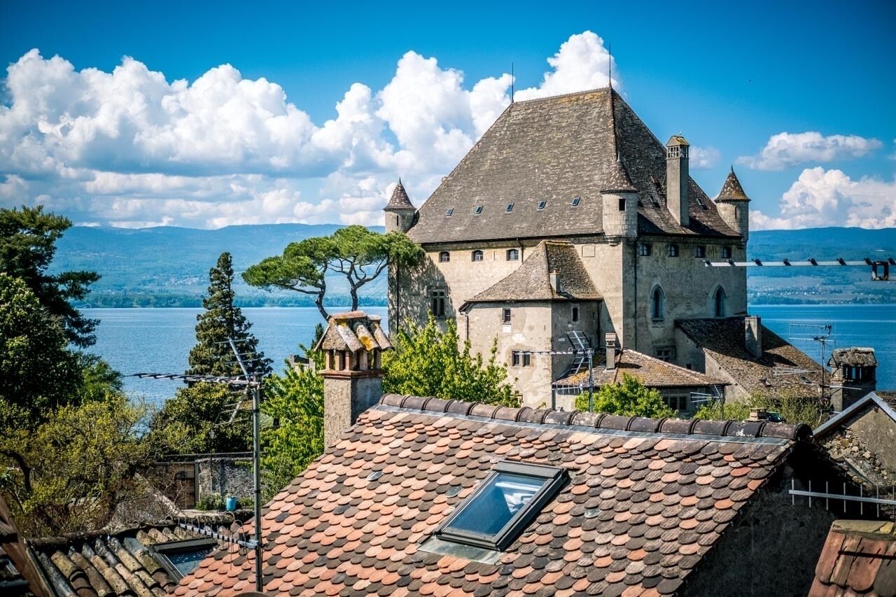 Un edificio histórico de piedra con un techo empinado y varias chimeneas, que domina un cuerpo de agua.