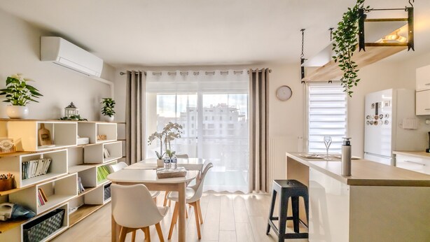 Una cocina moderna con armarios blancos, un comedor con mesa y sillas blancas, y vistas a edificios a través de cortinas translúcidas.