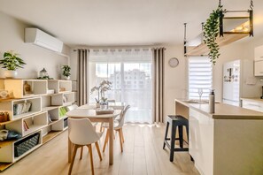Una cocina moderna con armarios blancos, un comedor con mesa y sillas blancas, y vistas a edificios a través de cortinas translúcidas.