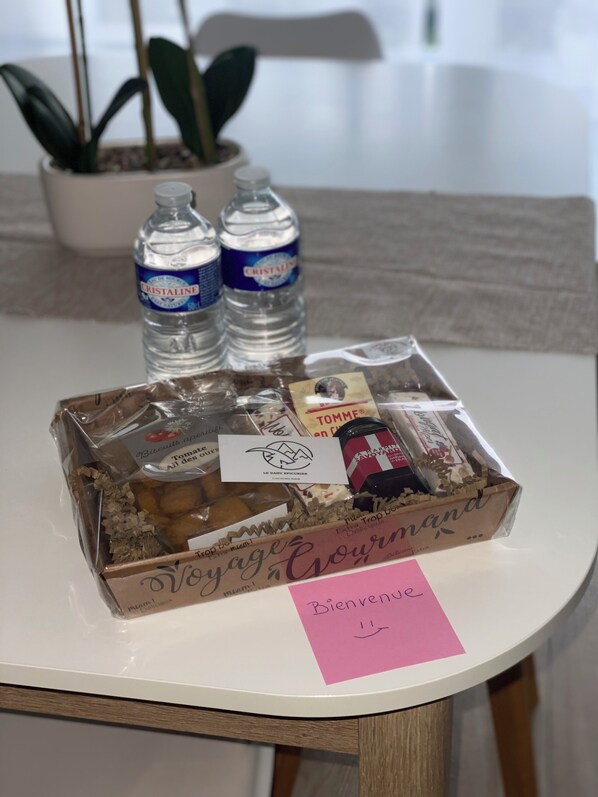 Una mesa con una caja de regalo etiquetada 'Voyage Gourmand', que contiene bombones, galletas y una botella de agua Cristaline.