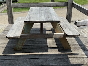 Una mesa de picnic de madera con diseño sencillo, sostenida por pilares de madera, ubicada sobre un piso de madera.