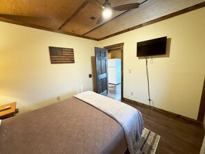 Una camera da letto con un letto, una ventola a soffitto, una televisione montata a parete e un'apertura che dà su un'altra stanza.